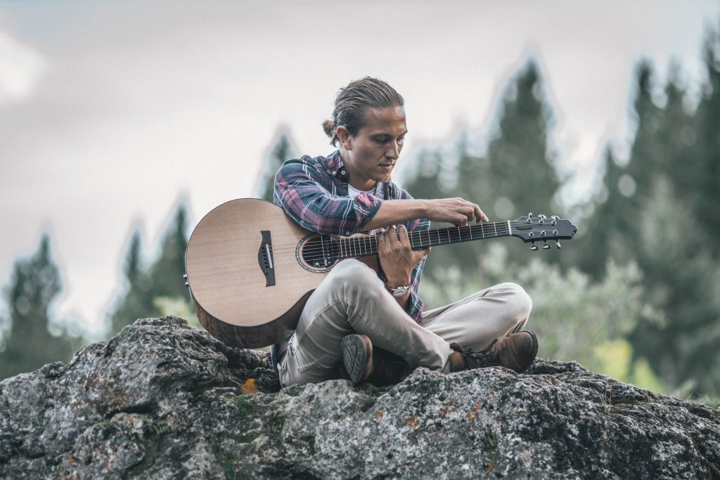 Callum graham sits on a lichen covered rock, playing guitar, the woods behind him of mountains and conifers. The swirling logo Callum uses for phoenix rising can be seen through the semi transparent background