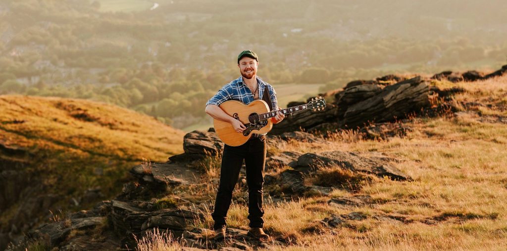 Jack Rutter standing in a landscape holding his guitar. Perhaps Yorkshire.